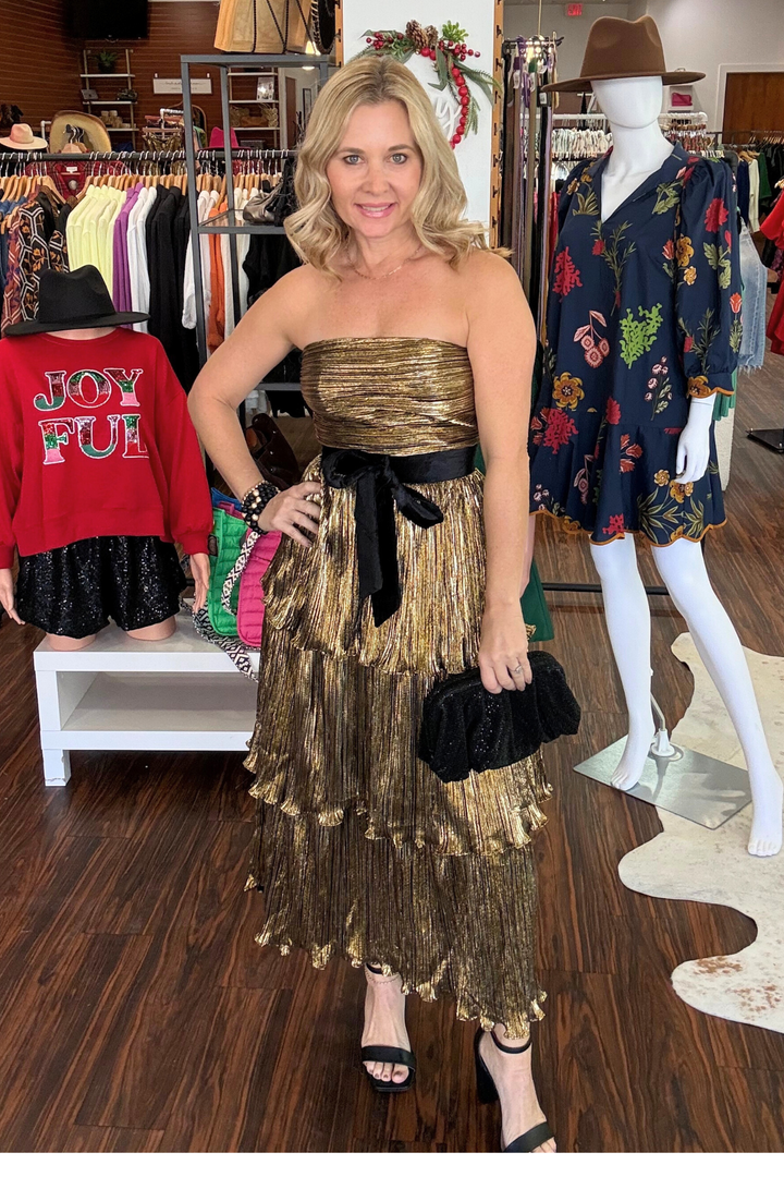 
Elevate your evening look with the Katia Gold Metallic Strapless Tiered Midi Dress. This sophisticated piece features a shimmering gold metallic finish that catches the light beautifully, paired with graceful tiered layers that create movement and dimension. The strapless silhouette offers a sleek, modern aesthetic, while the dramatic black velvet bow cinched at the waist adds a touch of refined elegance. 