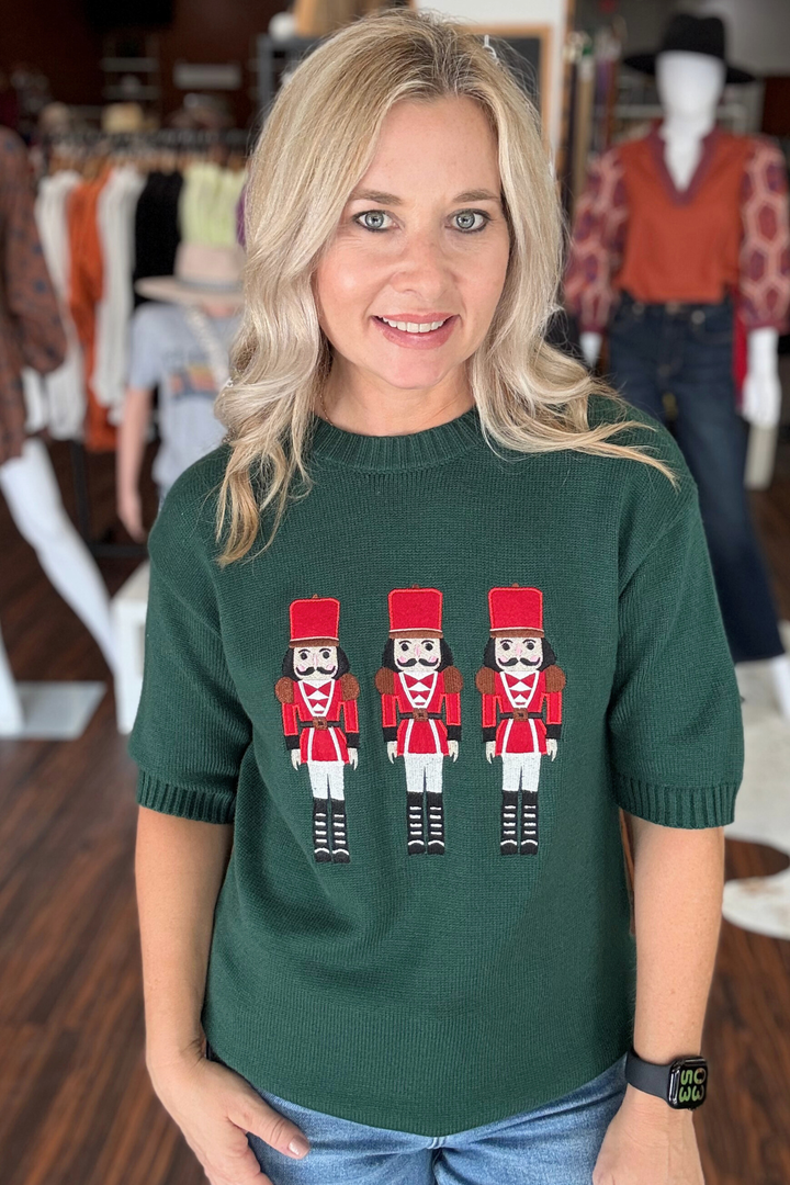 This hunter green short sleeve sweater features a modern design with three bold red nutcrackers across the front, adding a playful festive touch to your wardrobe. 